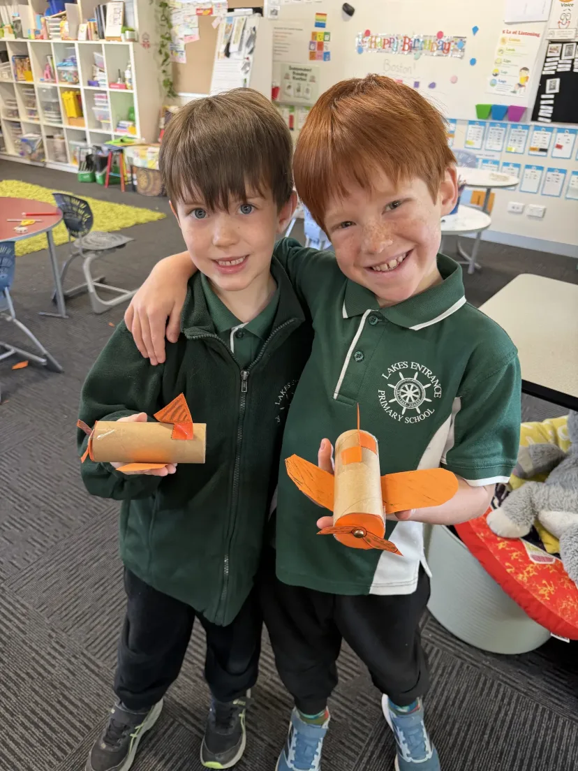 Two students with their arms round each other holding model planes they built